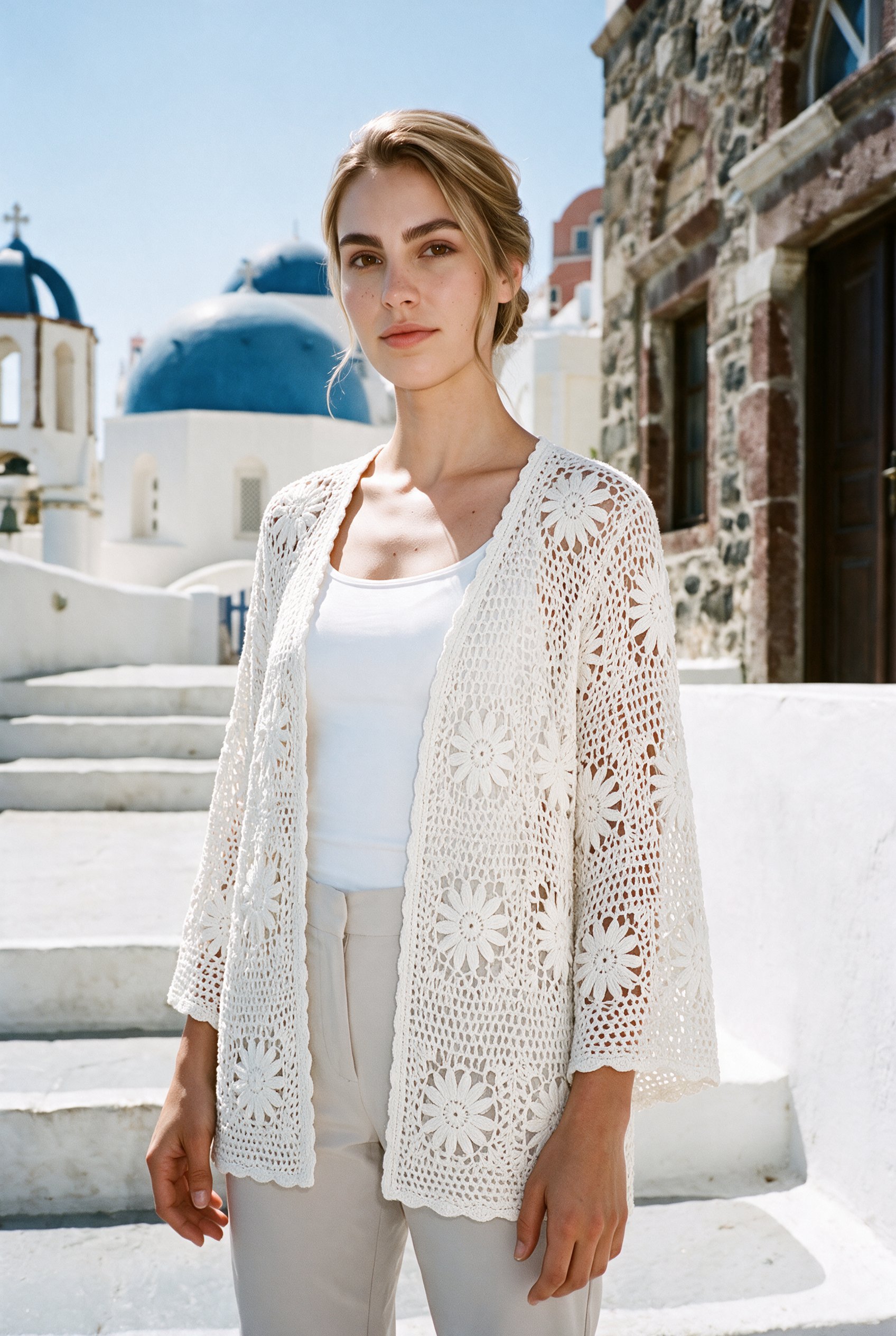 A 23-year-old Caucasian woman wears a white crochet cover-up in an upper-body, angled-front view with her arms relaxed and a neutral expression. White Santorini steps form the backdrop.