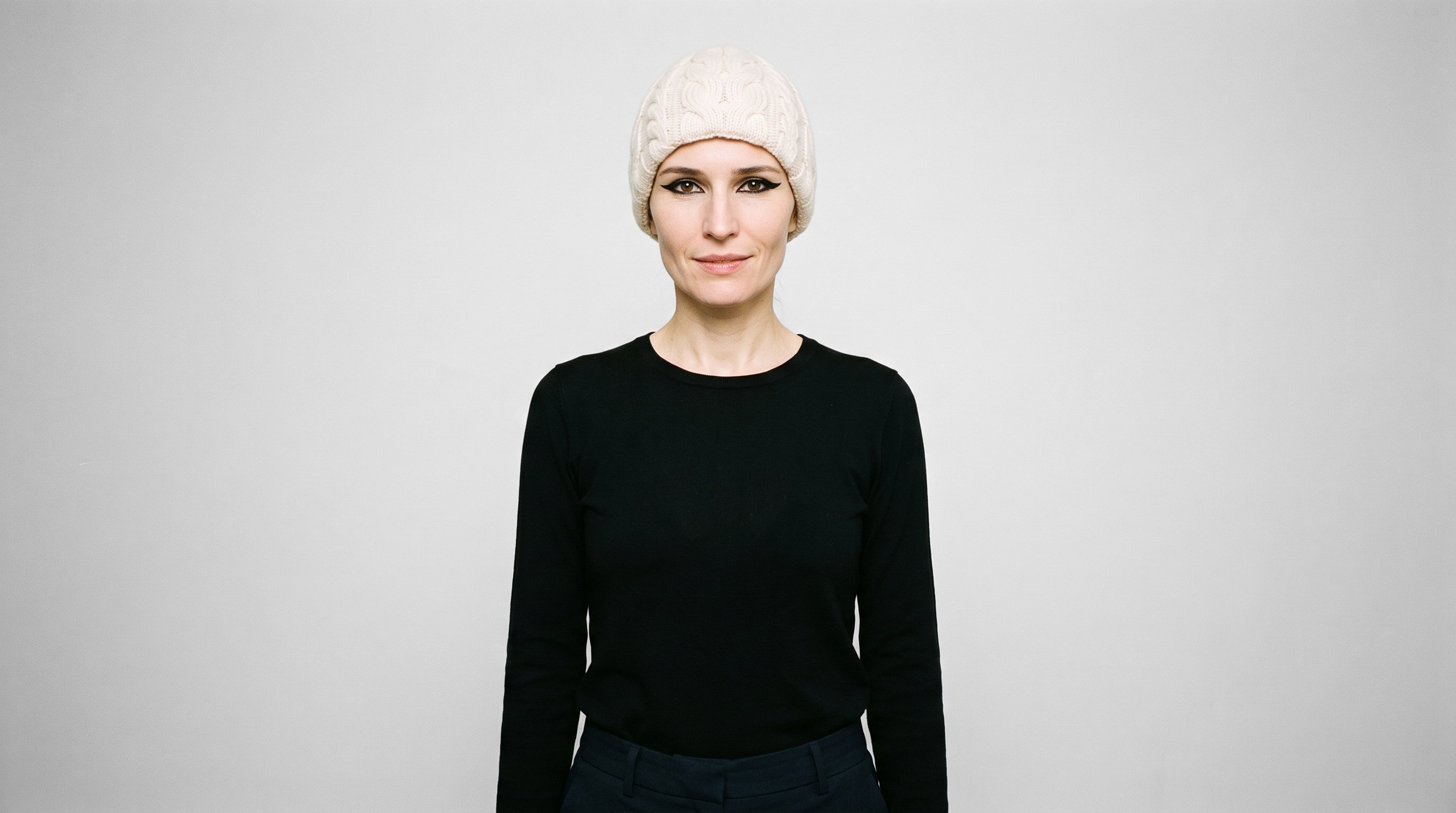 A 39-year-old Slavic woman wears a cream cable-knit beanie in an upper-body, front-view composition. Her arms are relaxed, and she has a confident expression against a cloud-white studio backdrop.