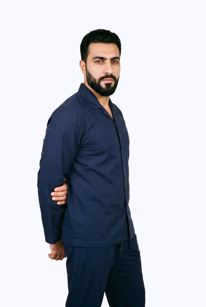 A 42-year-old Arab man wears a men’s navy cotton pyjama set, shown in a three-quarter-length front three-quarter view with one elbow positioned behind his back. His expression is soft, and a flat porcelain-colored backdrop frames the studio composition.