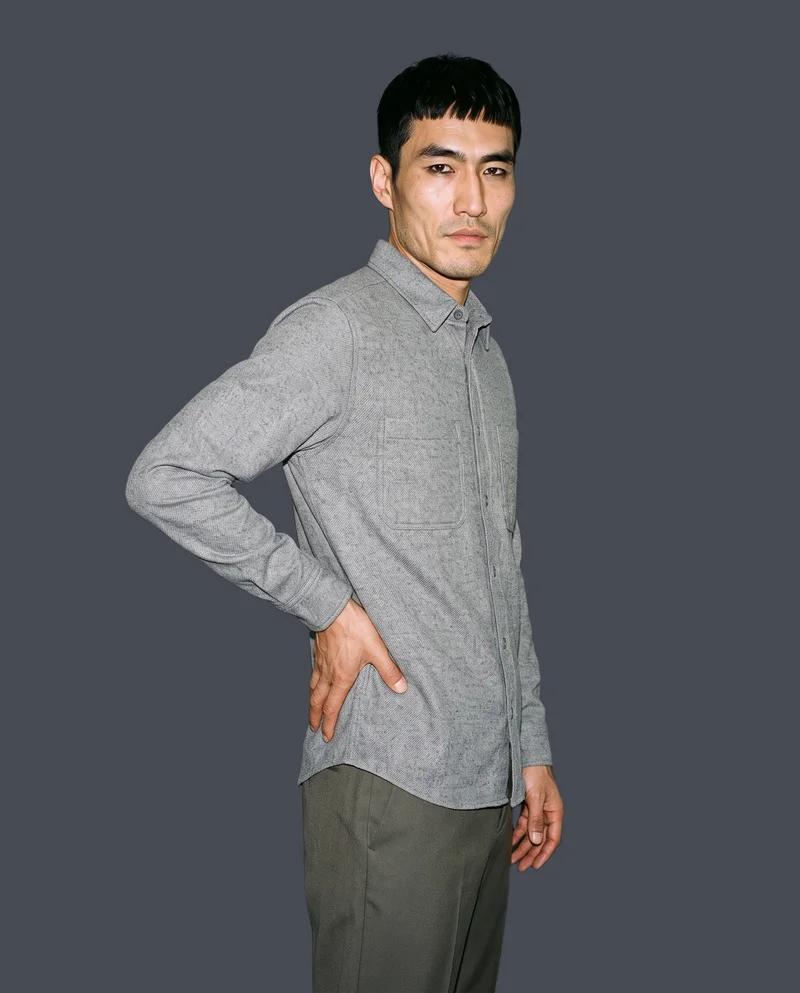 A 37-year-old Chinese male model wears a grey wool overshirt in a three-quarter-length view from a front three-quarter angle. He stands with one hand on his lower back and a neutral expression against a flat graphite backdrop.