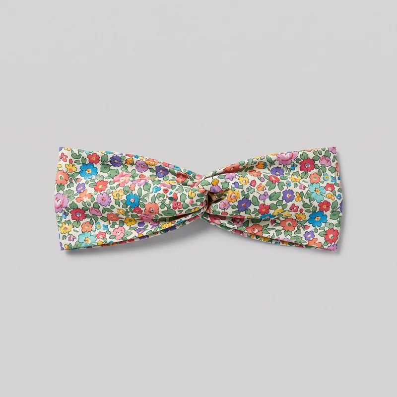 A floral fabric hair headband is laid flat, showing its full front-facing band and centered twisted knot. Overlapping folded panels extend horizontally from the twist, with gathered layered fabric adding volume at the center and straight-edged ends with folded seams. Scattered blossoms and leaves cover the exterior, with no visible logos or additional embellishments.
