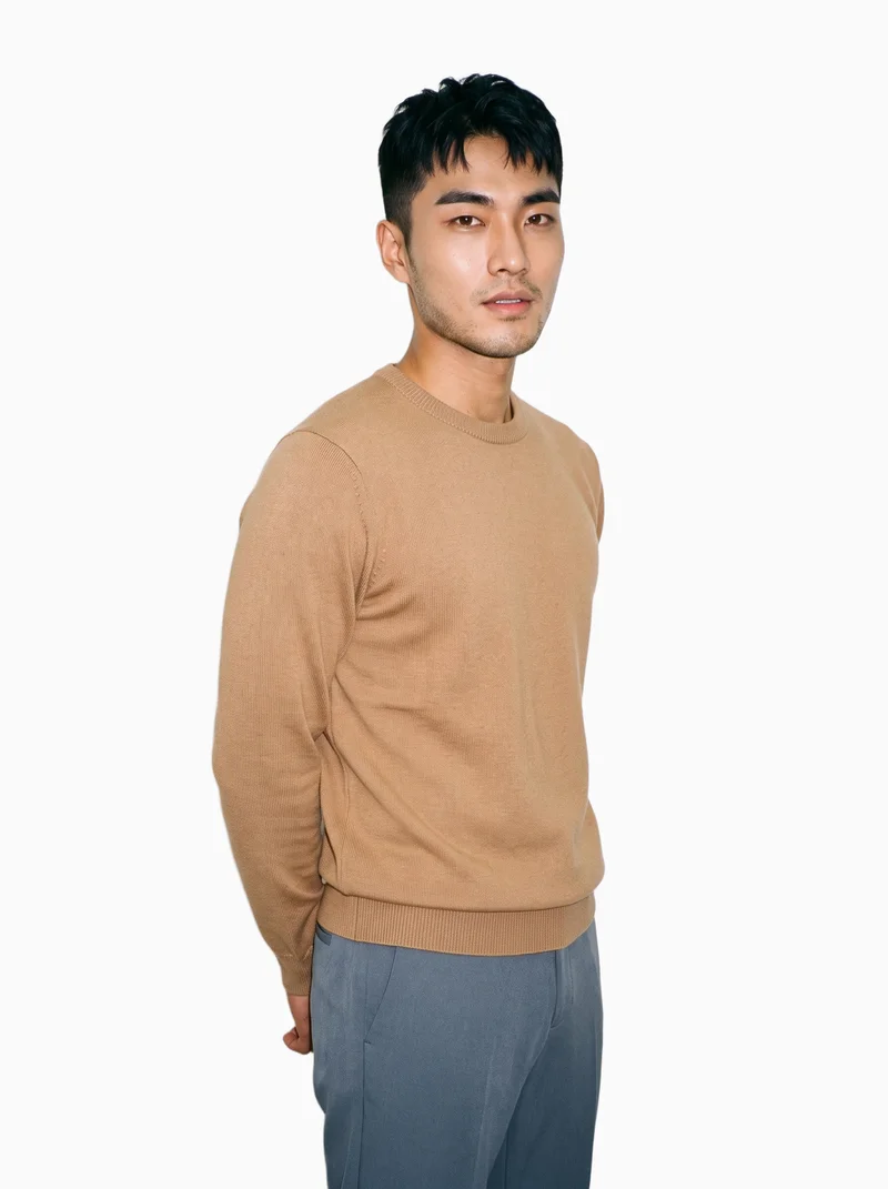 A 24-year-old Korean male model wears a camel knit pullover jumper, shown from a front three-quarter angle in a three-quarter-length frame with his hands behind his back and a soft facial expression. The flat-color Seasalt backdrop keeps the focus on the jumper and its overall silhouette.