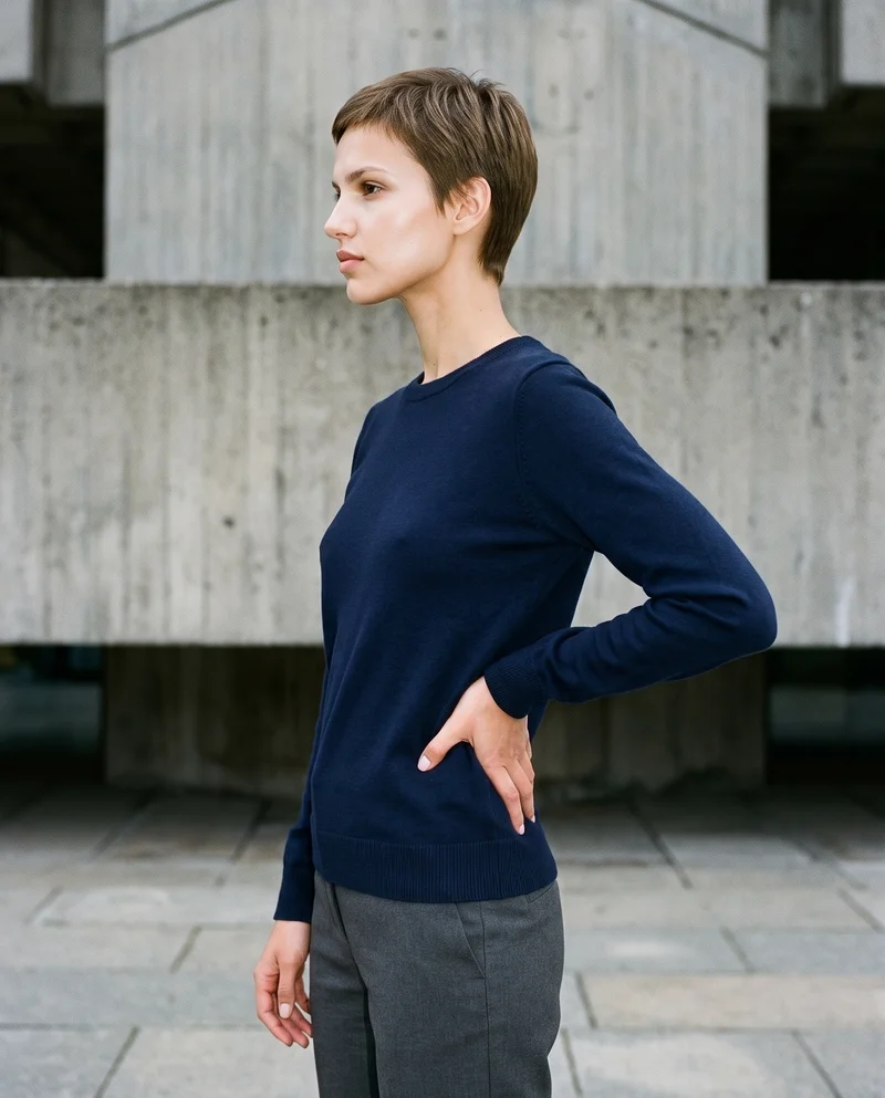 A 25-year-old Slavic woman wears a navy merino knit in a three-quarter-length side view, with one hand on her waist and a confident expression. A Brutalist Monument Plaza forms the backdrop.