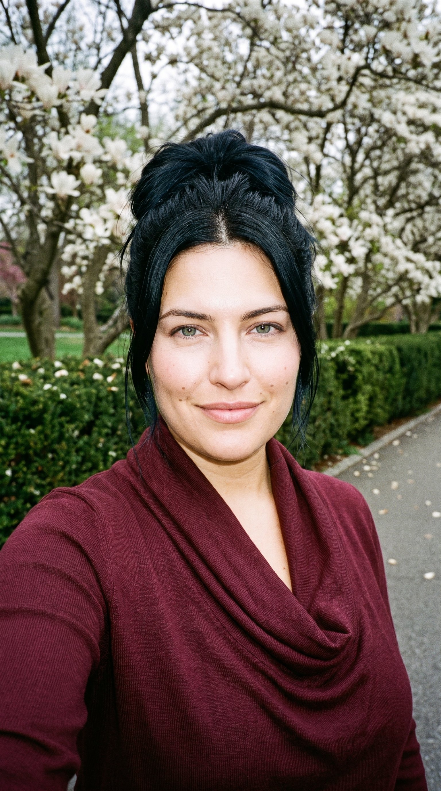 A 34-year-old Caucasian woman smiles at head level in a front-facing selfie while wearing a burgundy cowl-neck top. A magnolia tree park forms the backdrop.