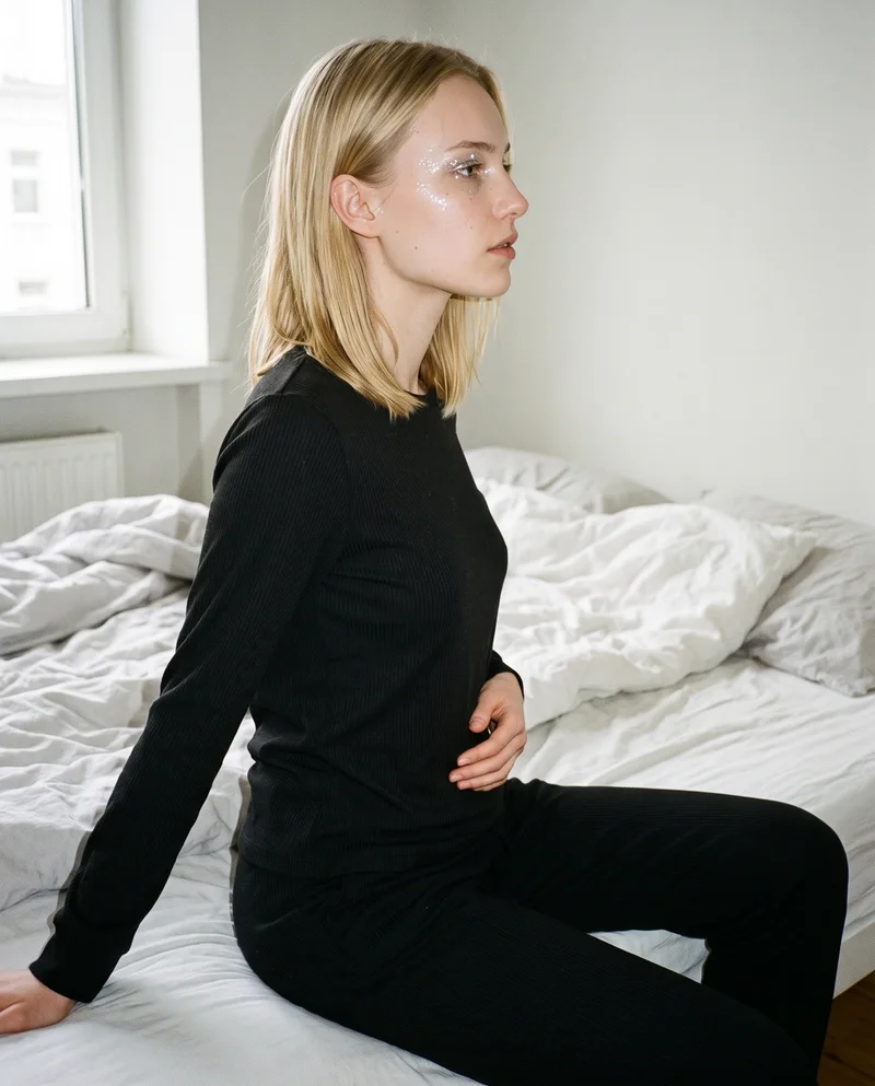 A 19-year-old Caucasian woman wears a women's black ribbed loungewear set in a three-quarter-length side view. She stands with one hip pushed outward and a serious expression, against a bedroom backdrop.
