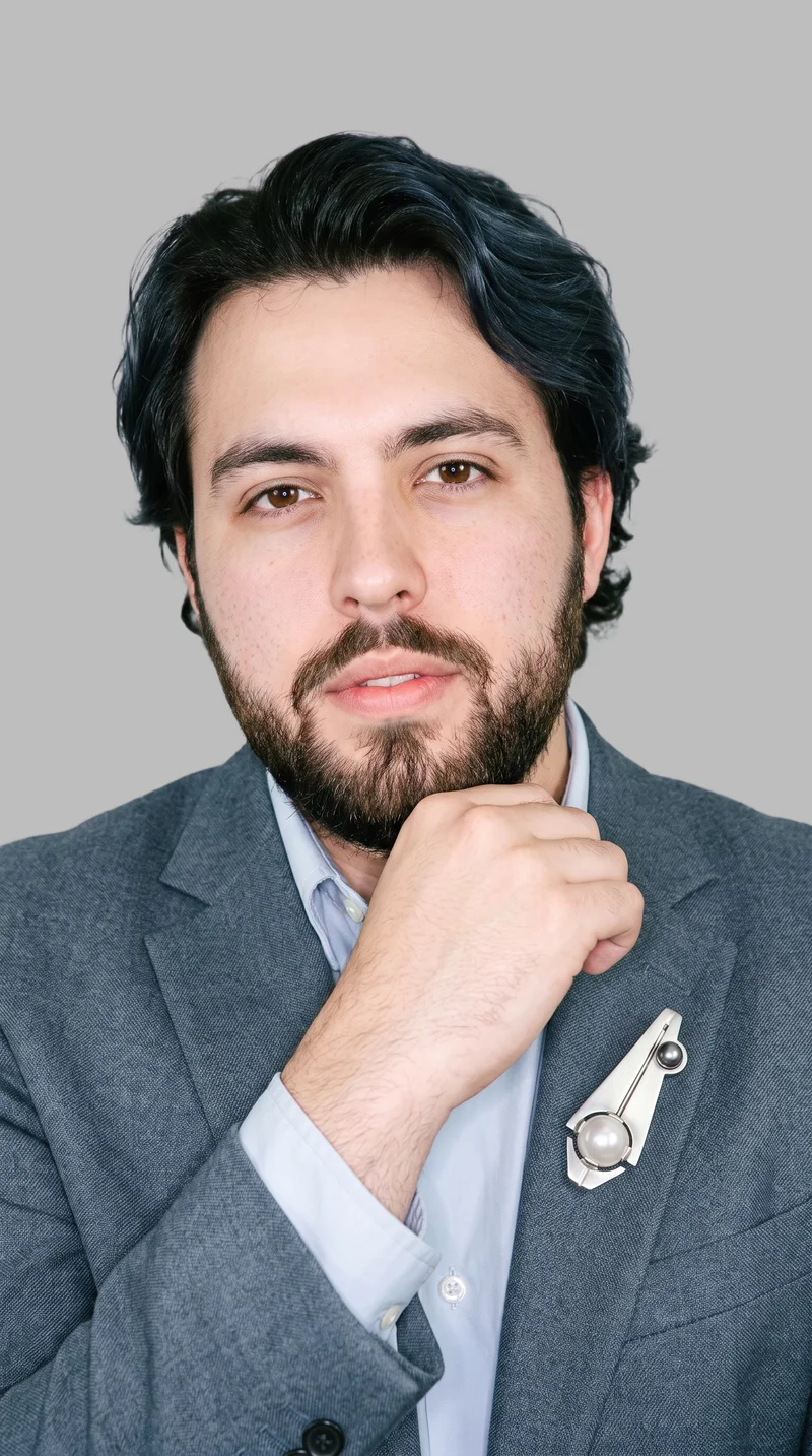A 25-year-old Caucasian man is shown in a front-view bust composition with a natural expression and his wrist held at his jaw, displaying a men’s pearl brooch. The image has a dove grey flat-color backdrop.