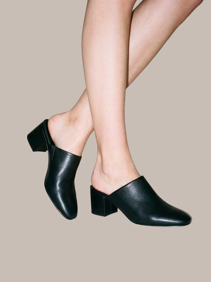 A pair of women's heeled black mules shown from an angled front view, with one heel lifted. Both feet are framed against a flat greige backdrop.