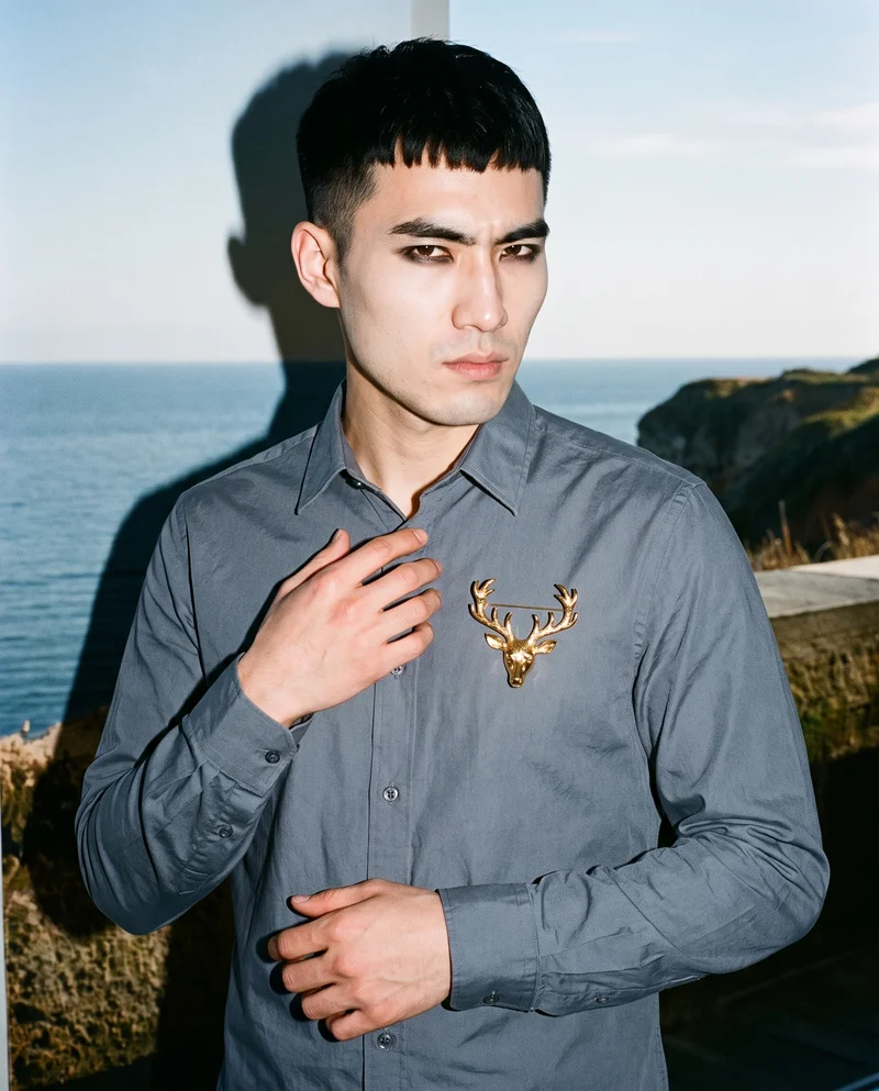 A 24-year-old Chinese man is shown from the upper body at a front three-quarter angle, wearing a gold brooch with his fingers held at his chest. He has an intense facial expression, with an Amalfi Coast terrace backdrop.