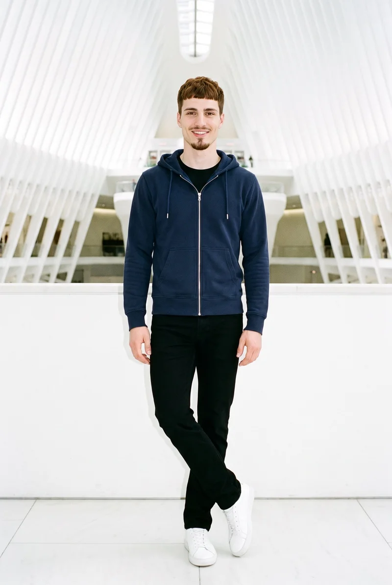 A 25-year-old Caucasian man wears a navy zip-up hoodie in a full-body front view, standing with his ankles crossed and a natural facial expression. A contemporary art white atrium forms the backdrop.