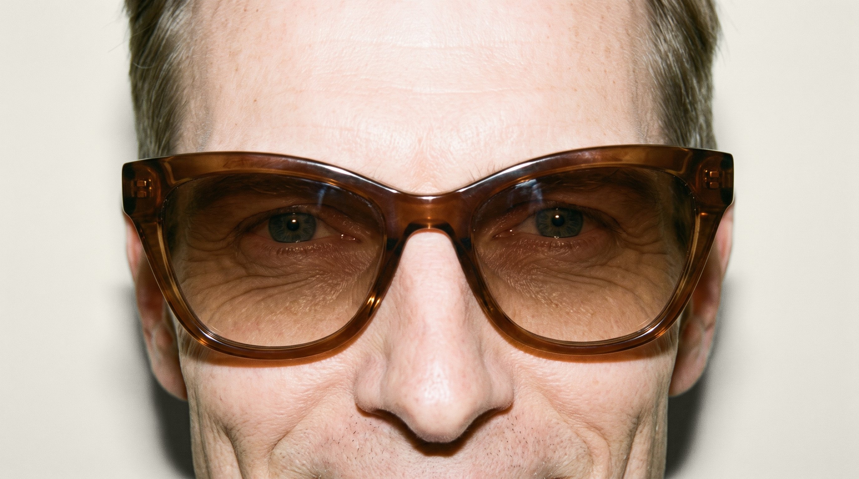 Brown cat-eye sunglasses shown from the front at eye level against an alabaster studio-wall backdrop.