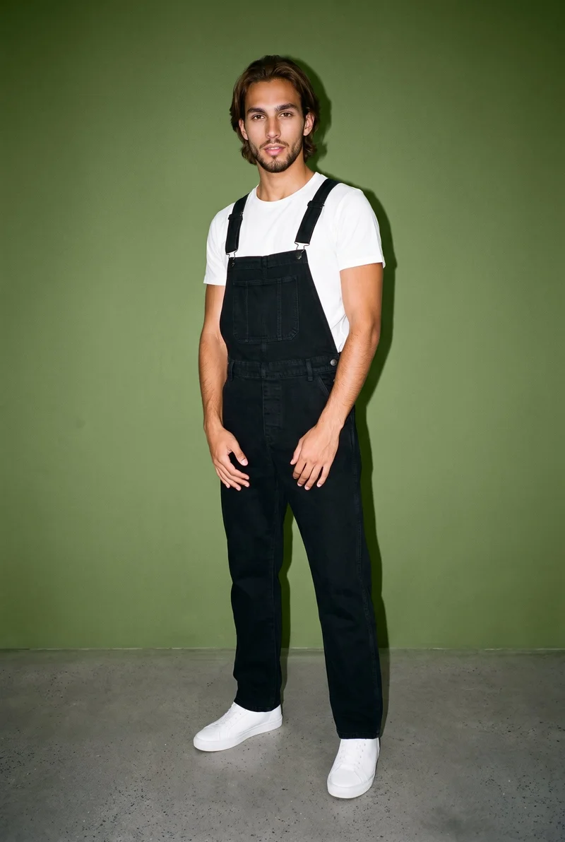 A 22-year-old Hispanic male model wears black denim dungarees in a full-body, angled-front view with his hands positioned in front. The olive grove backdrop is presented as a studio wall.