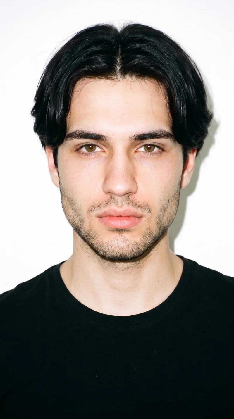 An 18-year-old Slavic male with fair skin and a neutral undertone has a triangle-shaped face, with a wide jaw narrowing toward the forehead and emphasis on the lower face. His eyebrows are thin, and his hazel eyes contain green-gold flecks. He has full lips and black hair divided at the centre, falling evenly to both sides. Dense heavy stubble forms a dark, defined layer across the beard area.