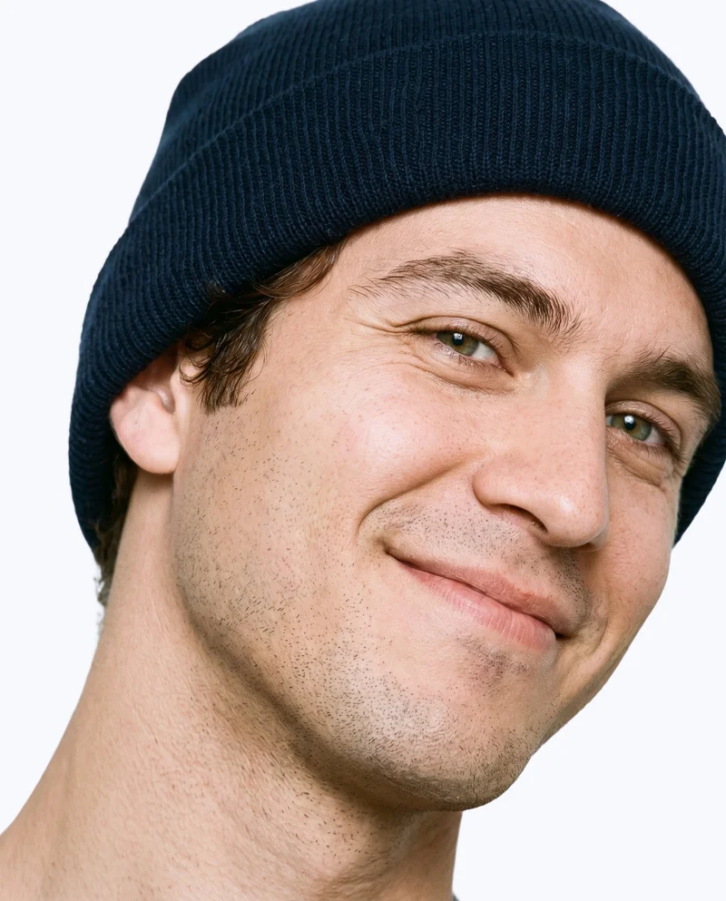 A 27-year-old Hispanic man wears a navy beanie, shown in an angled front view with his head tilted and a natural expression. The subject is framed from the head against a flat porcelain-colored backdrop.