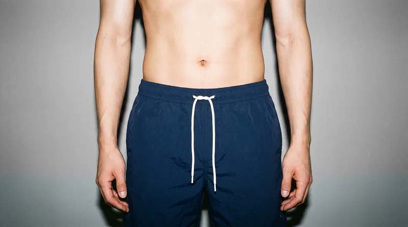 Navy swim shorts shown from the front on a male model, framed from the hips with the hips level. The shorts are presented against a pebble grey studio-wall backdrop.