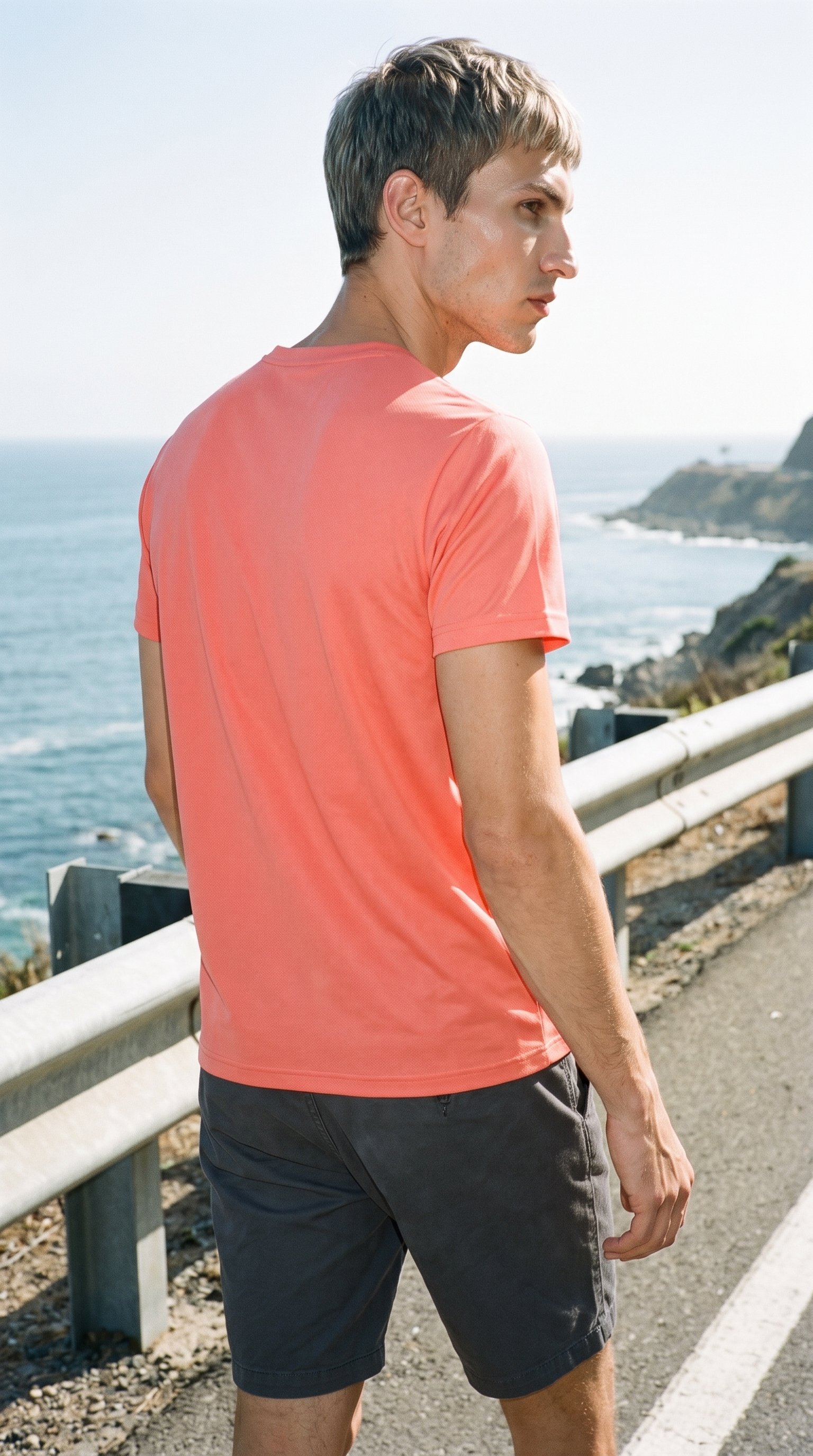 A 27-year-old Slavic man wearing a coral seamless performance top is shown from an angled rear view in a three-quarter-length frame while walking. The Pacific Coast Highway appears as the backdrop.
