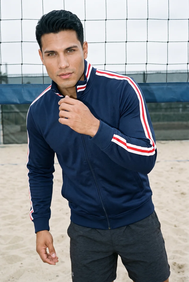 A 27-year-old Hispanic man wears a men's navy retro track jacket in an upper-body front view. He poses with one hand at his chin and a confident expression against a beach volleyball court backdrop.