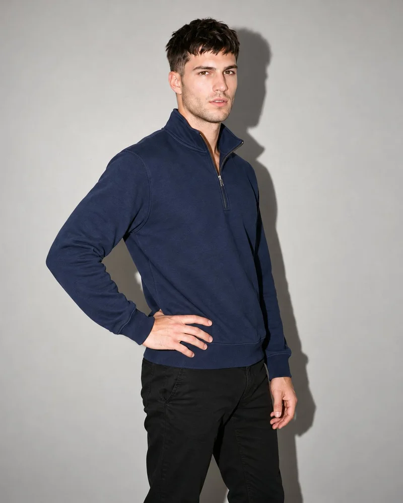 A 21-year-old Caucasian male model wears a men's navy quarter-zip, shown from an angled front view in a three-quarter-length frame with one hand on his hip. The image uses a Couture Grey studio-wall backdrop and an editorial facial expression.
