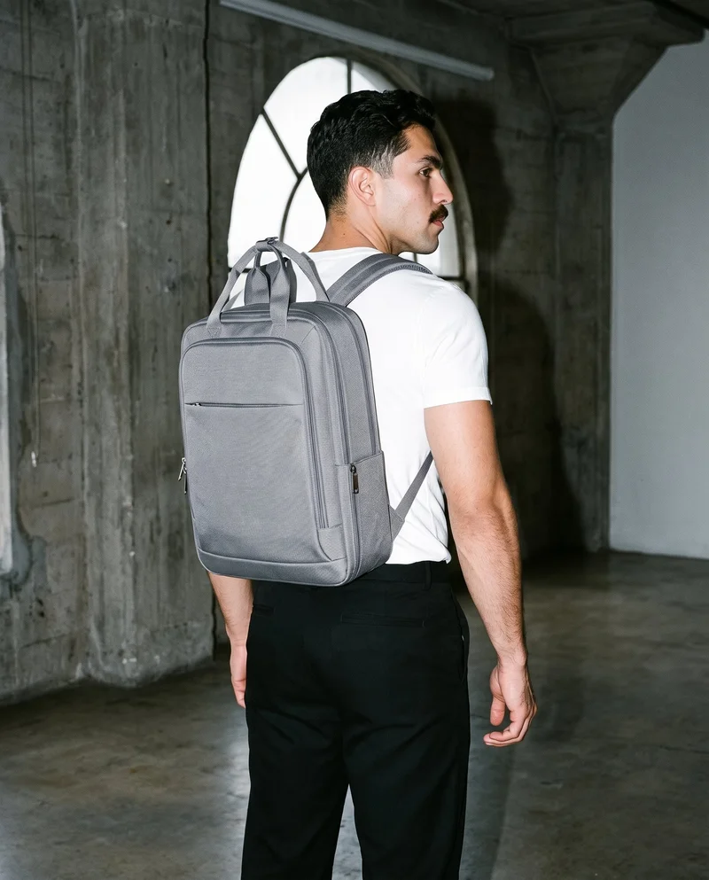 A 24-year-old Hispanic man is shown in a three-quarter-length frame carrying a grey laptop backpack on his back, viewed from an angle behind him. Massive loft windows with golden light form the backdrop.