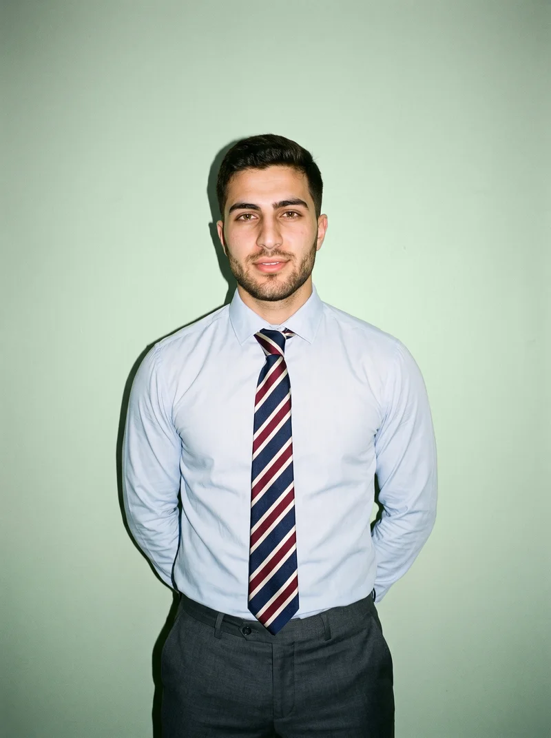 A 19-year-old Arab man is shown from the front in an upper-body view wearing a striped tie, with his hands behind his back and a natural facial expression. A pistachio studio-wall backdrop fills the background.