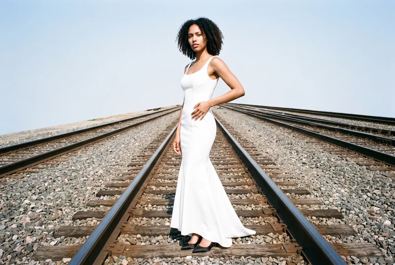 A 22-year-old African woman wears a mermaid satin wedding dress in an environmental front three-quarter view, standing with one foot forward and a serious expression. Train tracks form the backdrop.