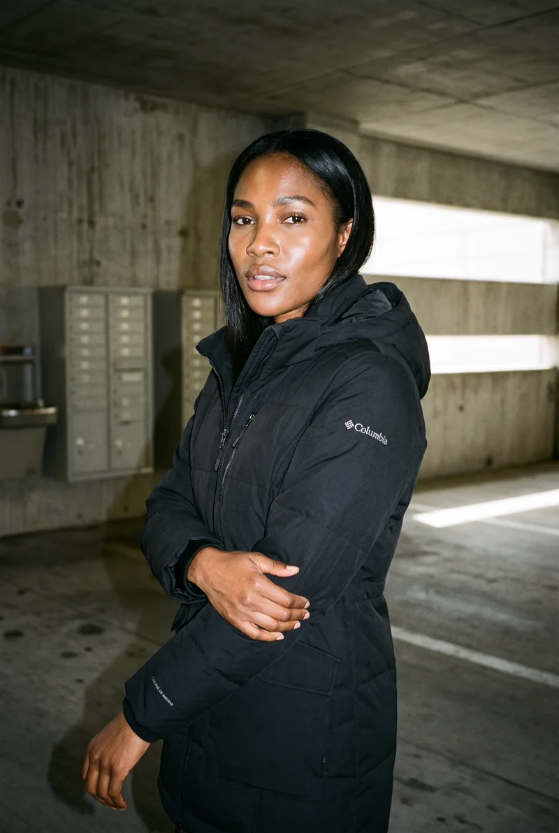 A 23-year-old African woman is shown from the upper body in a front three-quarter view, wearing a black parka and touching her wrist with a soft facial expression. A concrete parking garage serves as the backdrop.