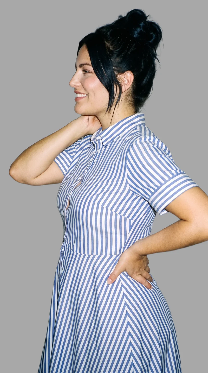 A 34-year-old Caucasian woman is shown from the upper body in side view, wearing a striped shirt dress with one hand behind her neck and smiling. A flat pebble grey backdrop frames the studio-cutout composition.