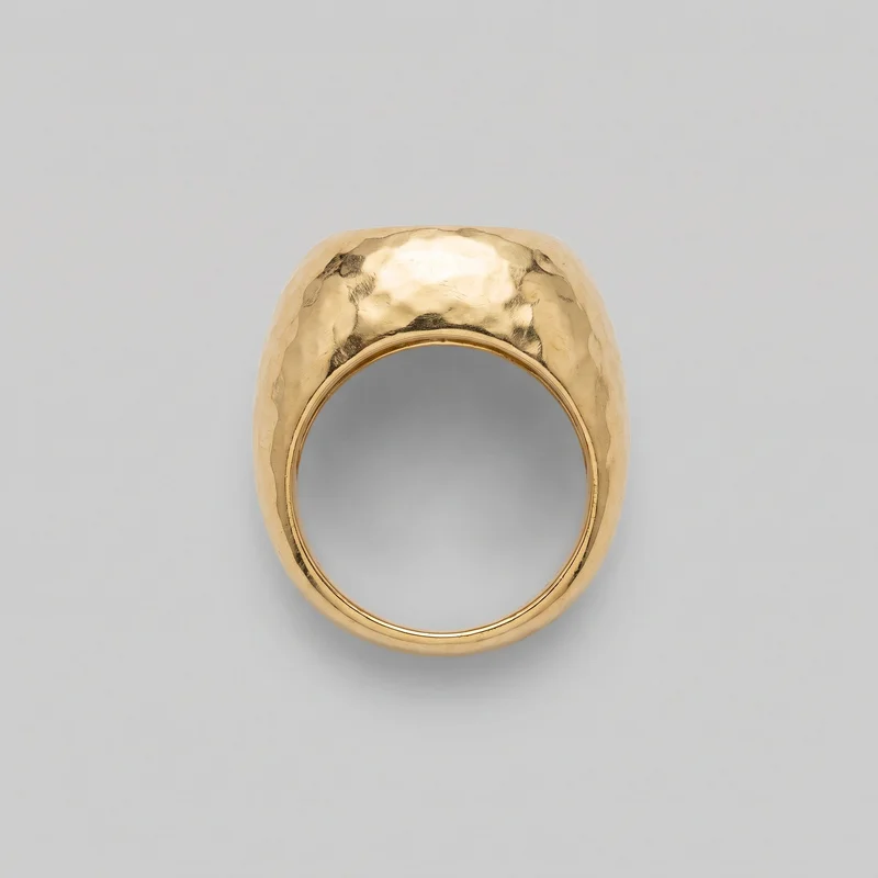 A chunky gold ring stands upright in a front view, showing its wide continuous band, oval finger opening, and raised domed top. The crown and shoulders have an irregular hammered, faceted surface, while the sides taper into the lower band with smooth rounded edges and a visible inner rim.