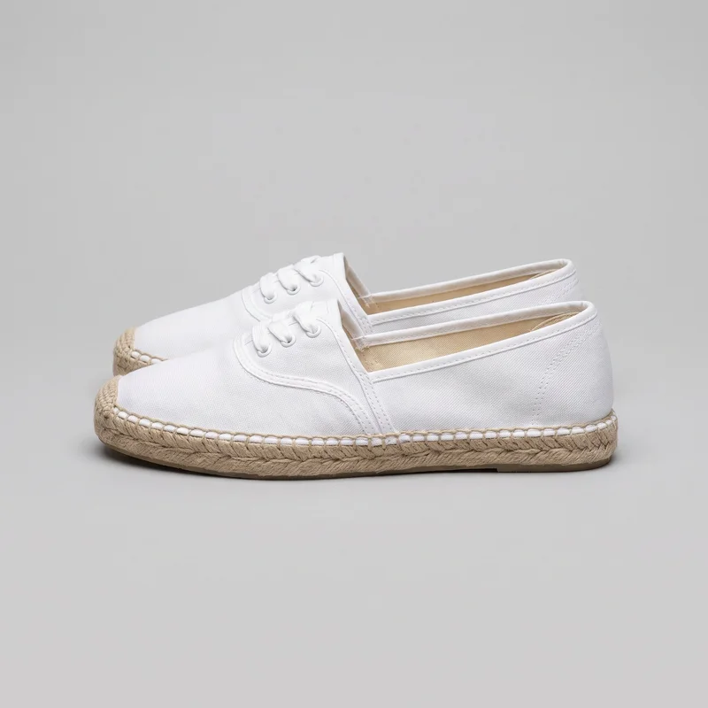 A pair of women's white low-profile lace-up espadrille flat shoes is shown from a side-front angle. The shoes have closed rounded toes, low-cut openings, tied laces through two visible eyelet pairs on each shoe, stitched facing panels, bound toplines, vertical heel seams, and visible stitching along the sides and collars. Braided espadrille-style sole wraps with looped stitching surround the edges above a thin outsole layer.