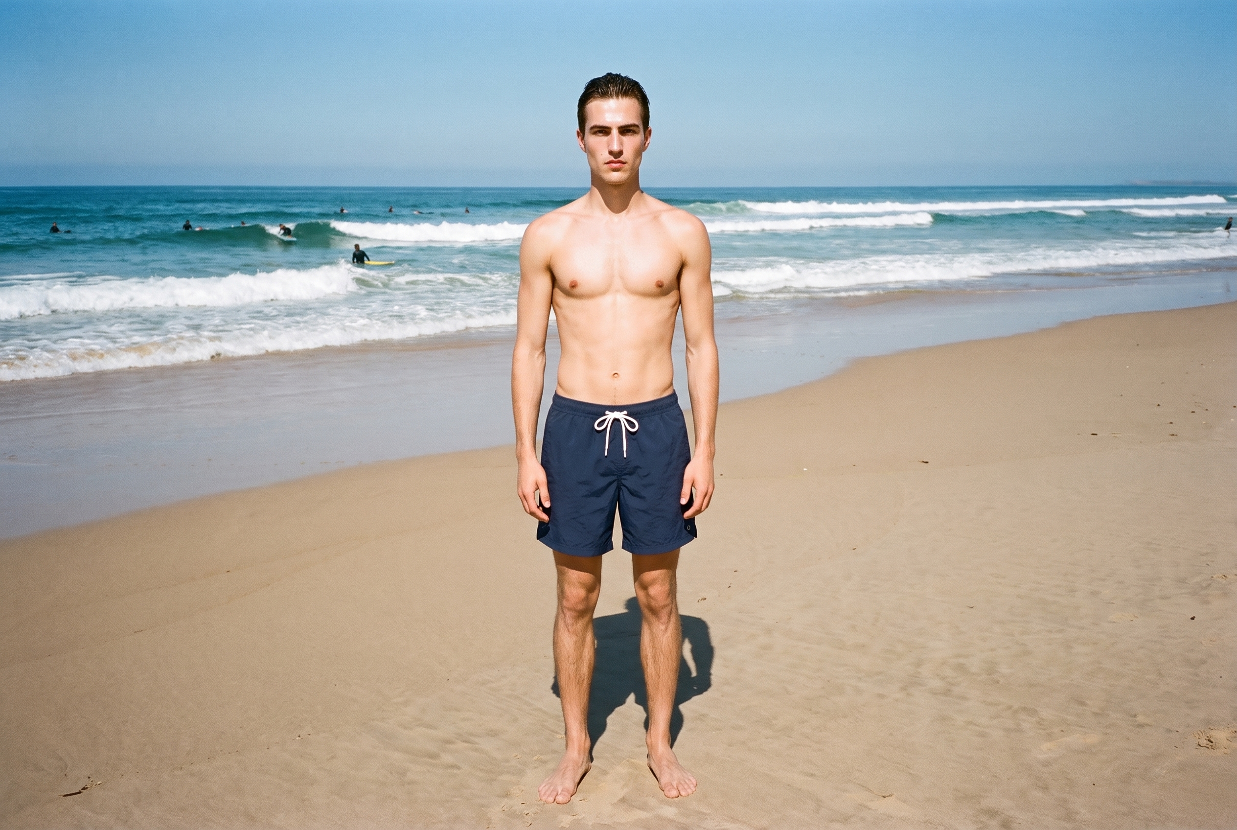 A 23-year-old Caucasian male model wears navy swim shorts in a front-facing environmental view, standing with his ankles crossed and a neutral expression. A surfing beach forms the backdrop.