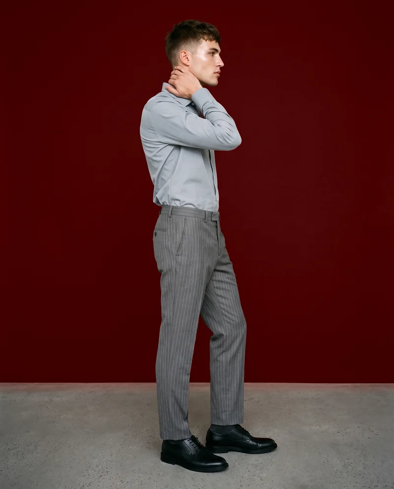 A 22-year-old Caucasian man is shown full body in side view wearing grey pinstripe suit trousers, with one hand behind his neck and a fierce facial expression. An oxblood studio-wall backdrop frames the composition.