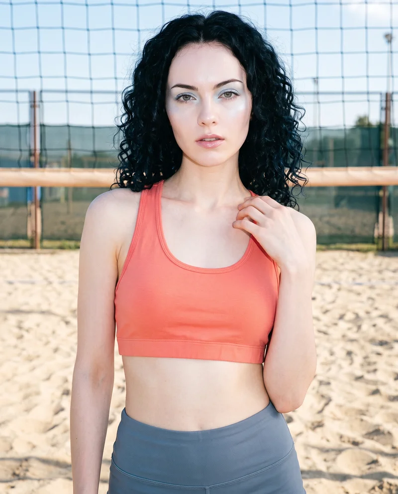 A 20-year-old Caucasian woman is shown from the upper body in a front-facing view, wearing a coral racerback sports bra with one hand at her collarbone and a natural expression. A beach volleyball court forms the backdrop.