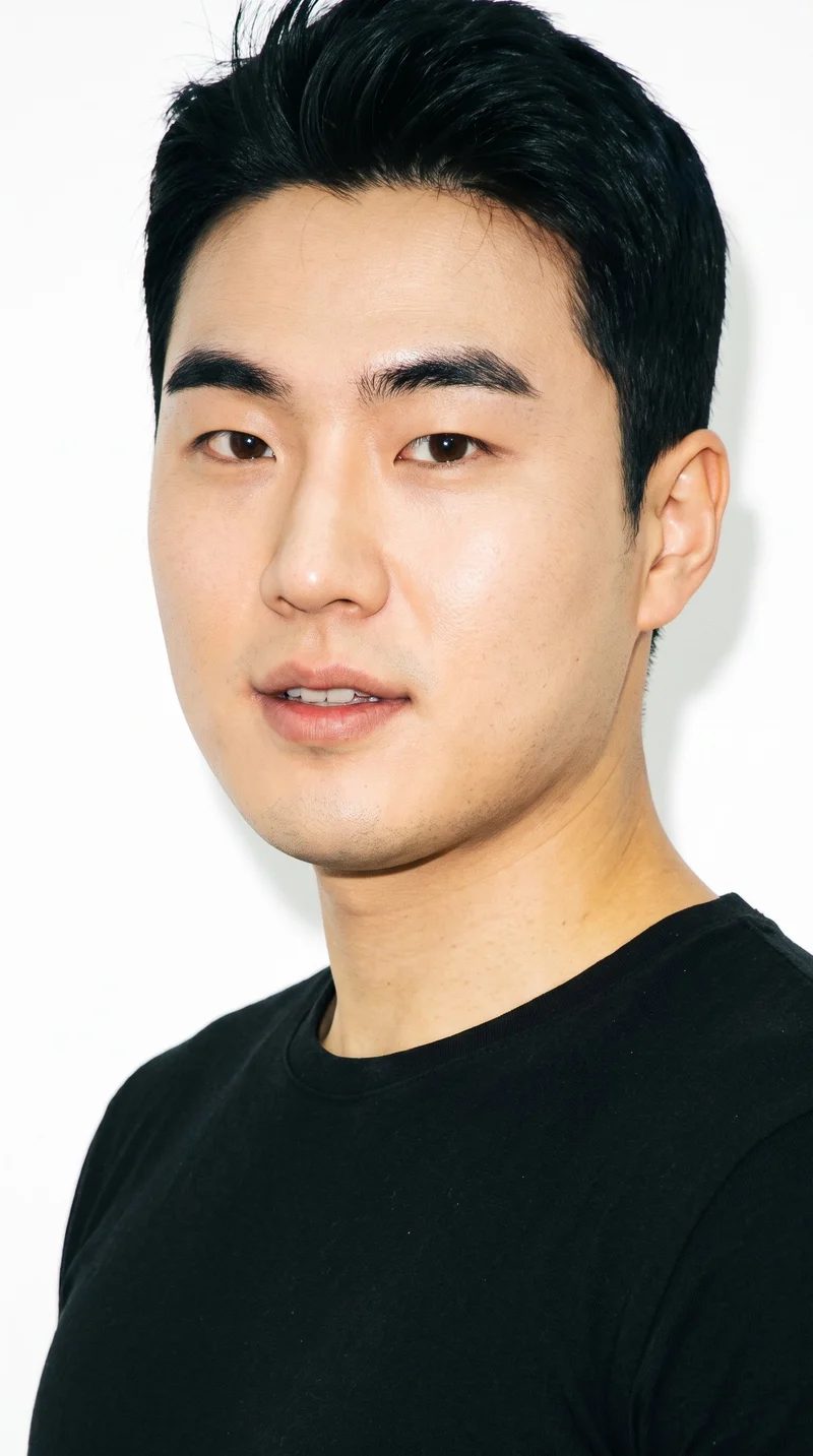 A 34-year-old Korean man with fair skin and a warm yellow undertone has a round face with soft, full contours and few angles. He has thick eyebrows, brown eyes, and thin lips. His black hair is styled in a short crew cut, tapered with slightly more length on top than at the sides, and he is clean-shaven.