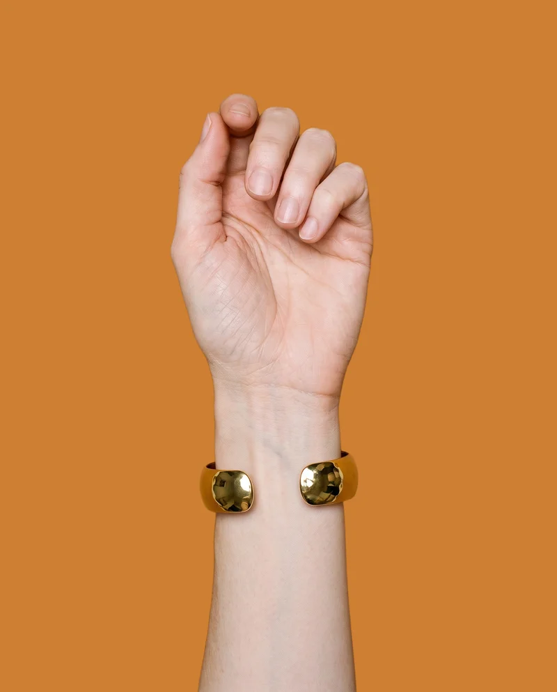 A woman's wrist and hand are shown from above with the knuckles facing forward, wearing a wide gold cuff. The bronze flat-color backdrop frames the hand and wrist.