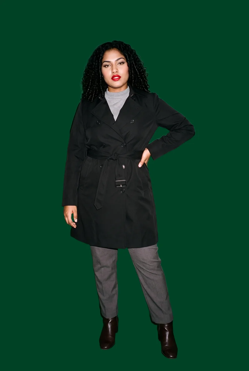 A 20-year-old Indian woman wears a women's black trench coat in a full-body front three-quarter view, with one hand on her hip and a curious expression. A flat British Racing green backdrop frames the studio-cutout composition.
