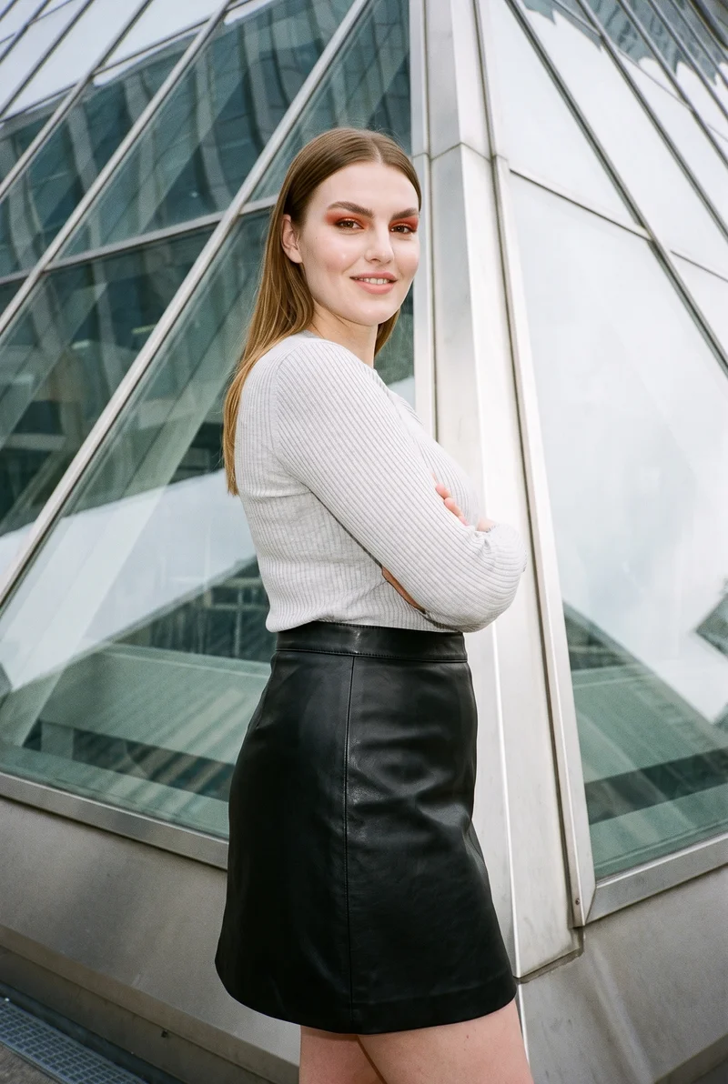 A smiling 25-year-old Caucasian woman wears a leather mini skirt in a three-quarter-length side view with her arms crossed. A pyramid building appears as a low-angle backdrop.