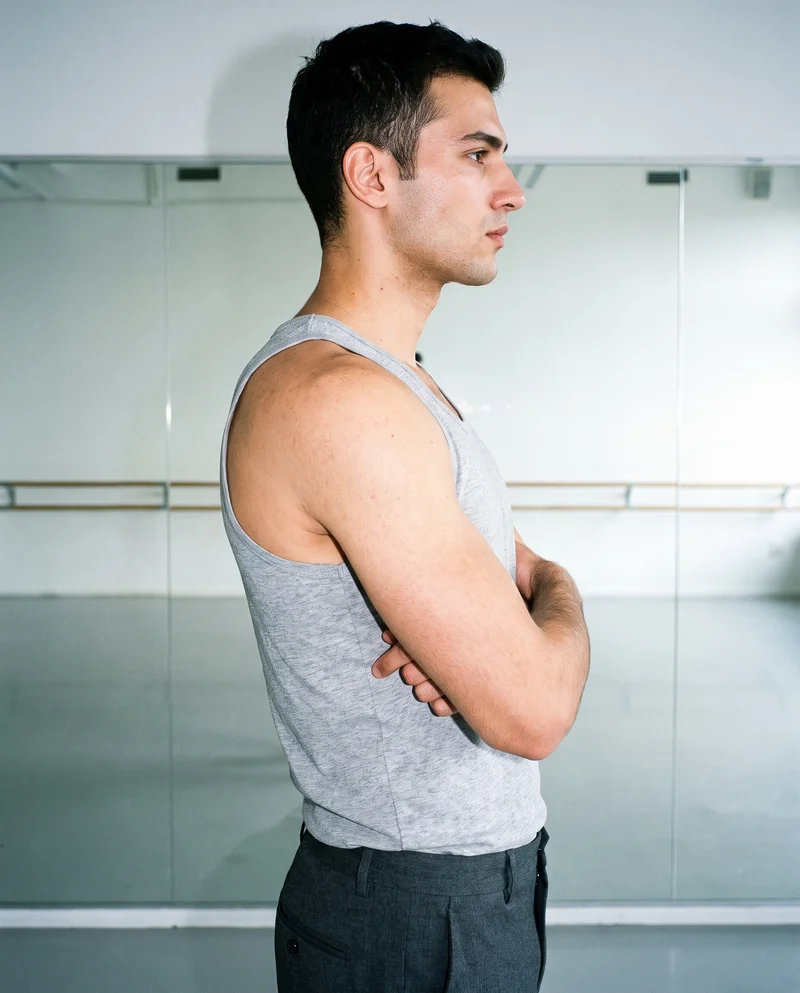 A 22-year-old Turkish male model wears a grey marl vest in an upper-body side view, standing with his arms crossed and a natural facial expression. A dance studio serves as the backdrop.