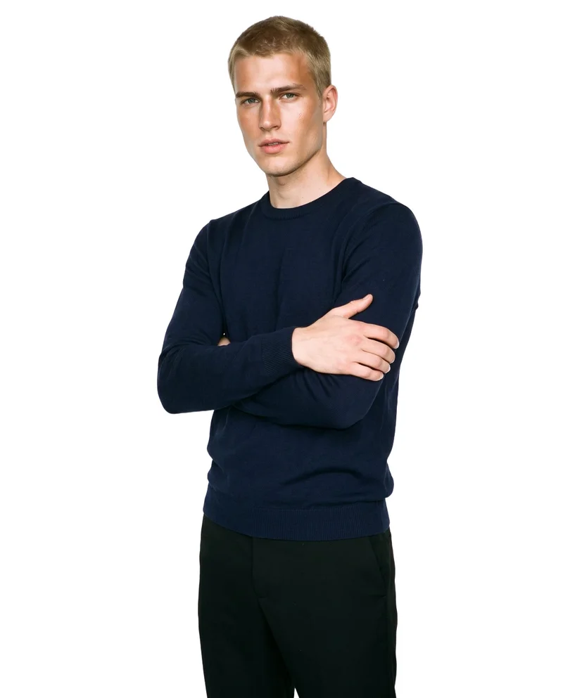 A 22-year-old Caucasian man wears a navy merino knit, shown from an angled front view in an upper-body frame with his arms crossed and a neutral expression. The garment is presented against a pure white backdrop.