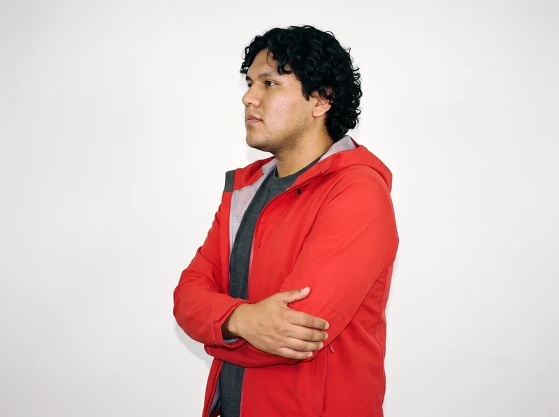 A 25-year-old Indigenous American man is shown from the upper body in side view wearing a red hardshell jacket. He holds himself in a self-hug pose with a neutral facial expression. A Chantilly Lace studio-wall backdrop fills the background.