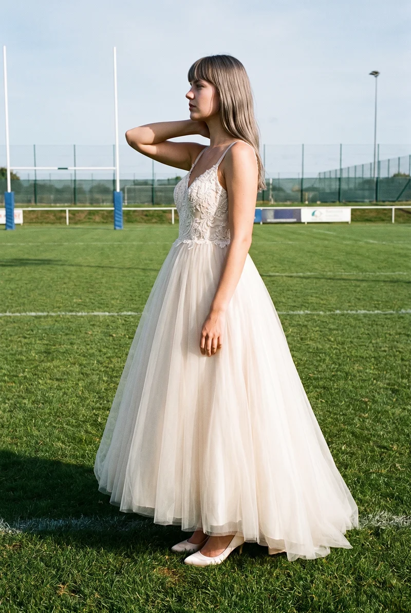 A 22-year-old Caucasian woman wears a champagne ballgown wedding dress in a full-body side-view composition. She poses with one hand behind her neck and a neutral expression, with a Rugby Field backdrop.