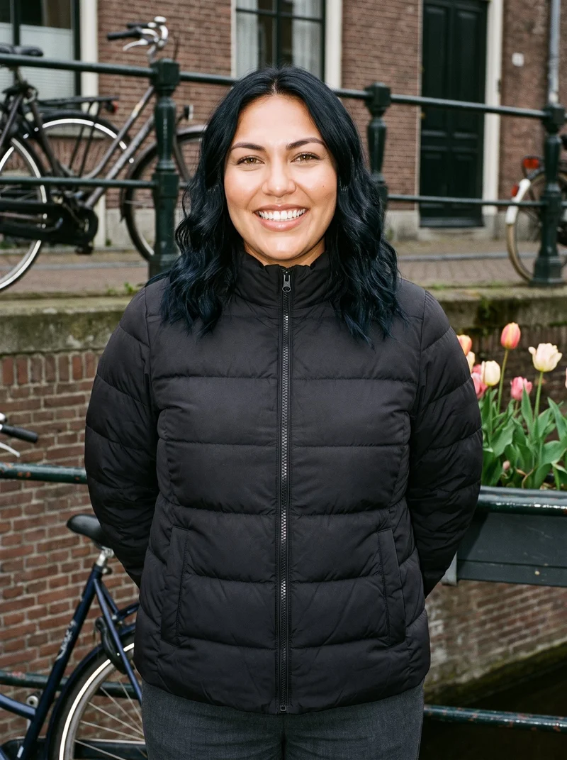 A 28-year-old Indigenous American woman wears a black puffer jacket in an upper-body, front-view composition. She stands with her hands behind her back and grins, with an Amsterdam canal house backdrop.