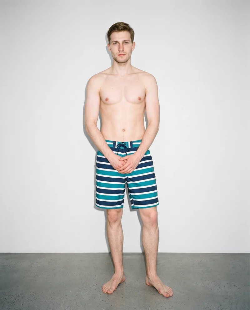 A 20-year-old Slavic man is shown full body from the front wearing striped board shorts, with his hands held in front and a neutral expression. A Fog Grey studio-wall backdrop fills the background.
