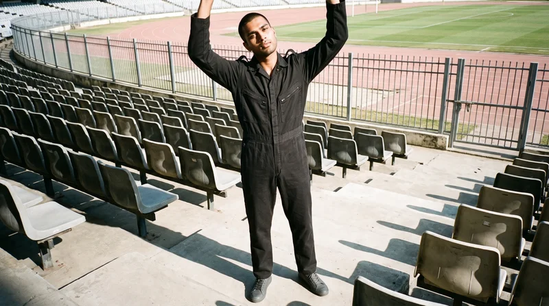 A 20-year-old Indian man wears a black boilersuit in an environmental frame, viewed from a front three-quarter angle with his arms overhead and a natural expression. Stadium seating forms the backdrop.