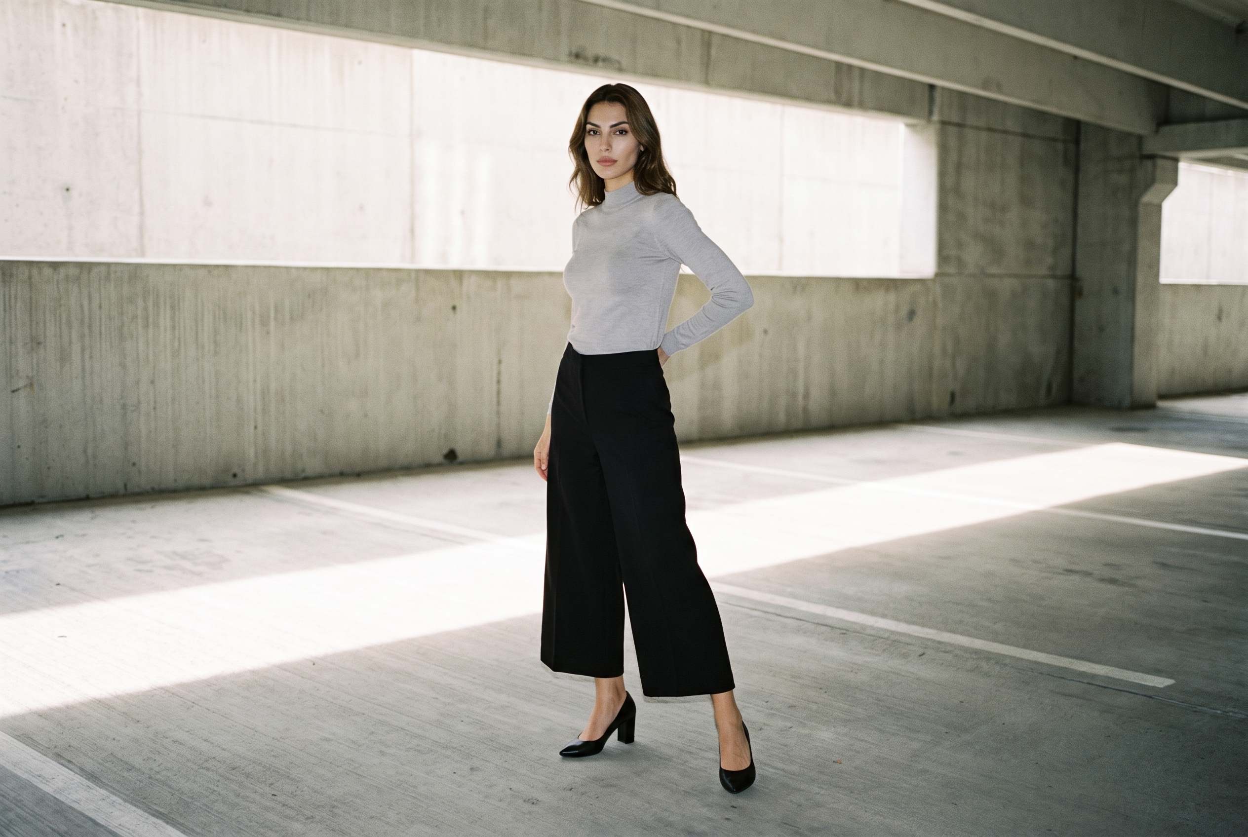 A 22-year-old Turkish woman wears black culottes in a full-body front three-quarter view, with one hand on her lower back and a neutral expression. A concrete parking garage serves as the backdrop.