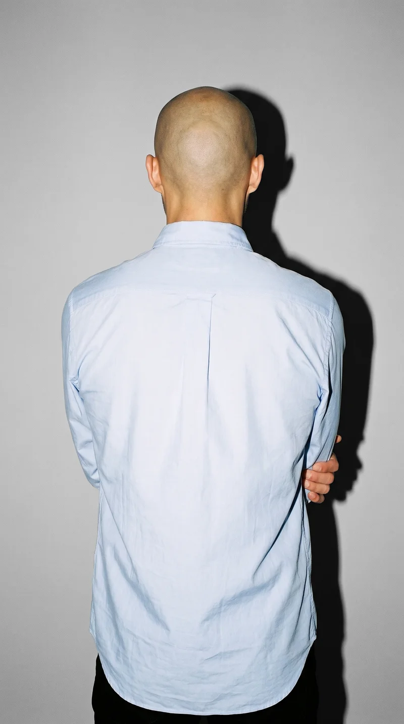 A 28-year-old Japanese man wears a light blue button-down shirt in an upper-body back view, holding his elbow. A dove grey studio-wall backdrop fills the background.