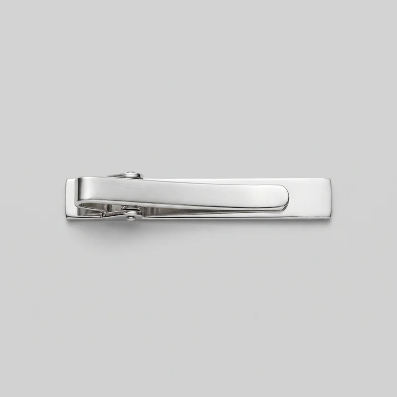 A complete silver tie bar clip is shown horizontally against a standalone product backdrop. The narrow rectangular accessory has a flat back plate, a rounded-corner front arm, a visible hinge pin, small round rivet-like caps, and an exposed tension spring mechanism near one end.
