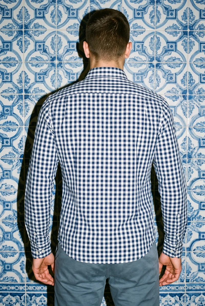 A 22-year-old Arab male model wears a navy gingham button-down shirt in an upper-body back view with a shoulder-drop pose. A Portuguese tile street forms the backdrop.