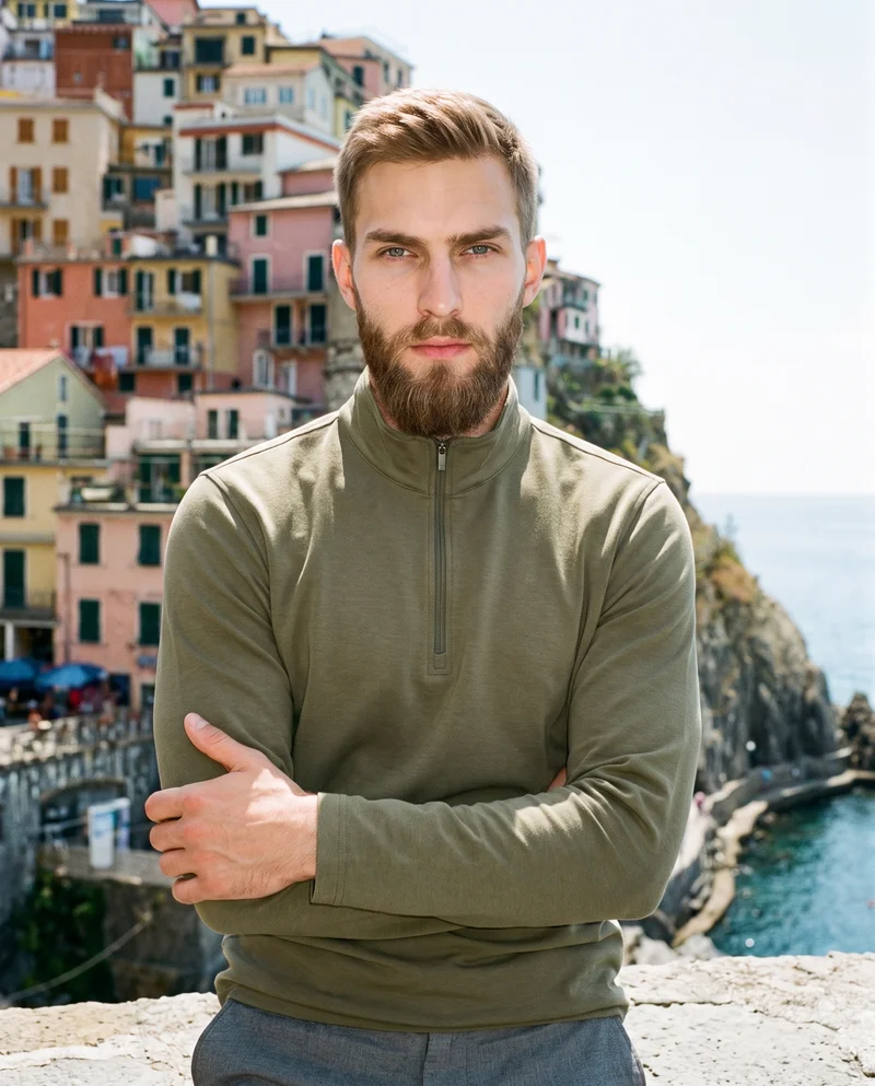 A 23-year-old Caucasian man wears a men's olive quarter-zip, shown from the upper body at a front three-quarter angle in a self-hug pose with a natural expression. A Cinque Terre cliffside serves as the backdrop.