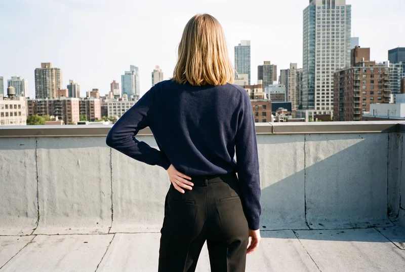 A 27-year-old Slavic woman wears a women's navy wool pullover jumper in a three-quarter-length rear view, with one hand on her hip. A warehouse rooftop skyline forms the backdrop.