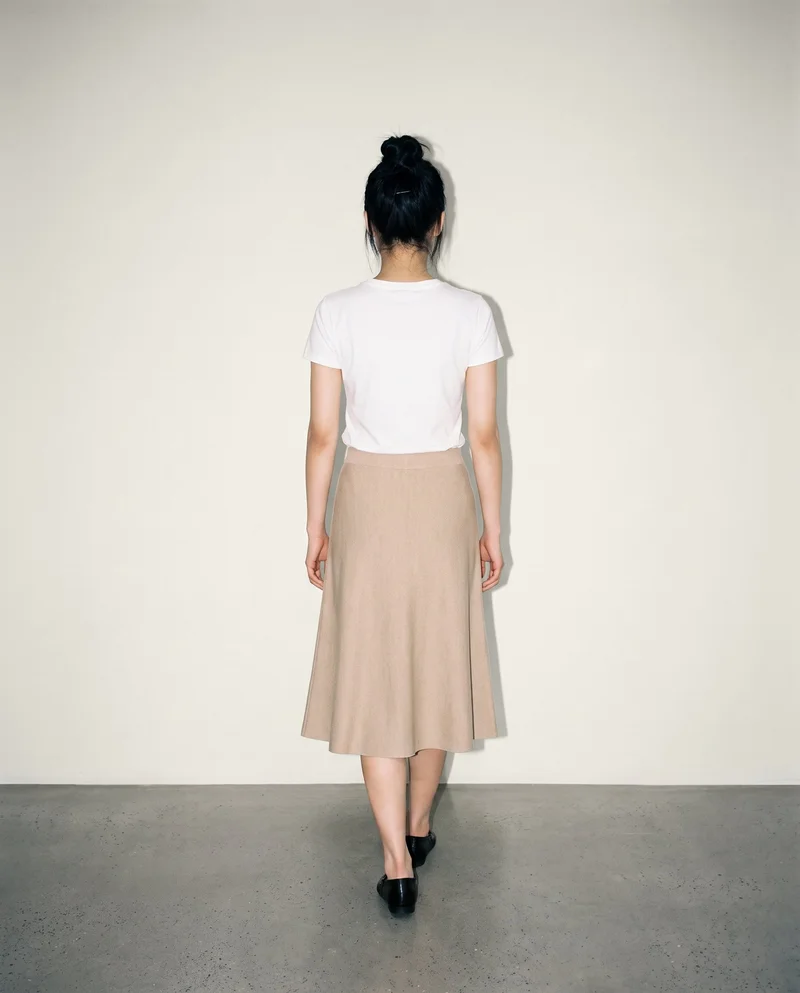 An 18-year-old Chinese woman is shown full body from the rear while walking in a beige knit midi skirt. The skirt is presented against an alabaster studio-wall backdrop.