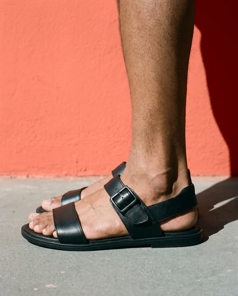 Black leather sandals shown from the side at ankle level against a terra cotta studio-wall backdrop.